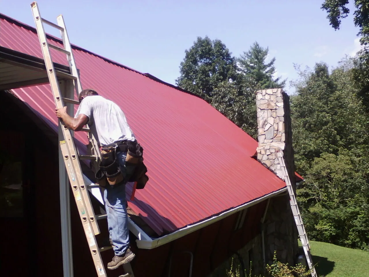 Roofer on a ladder beside a red metal roof and stone chimney — Soffit & Fascia Repair by Roofing Brothers in Bowersville